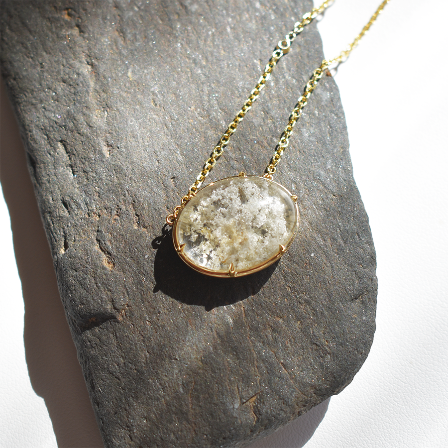 An oval quartz charm with a desert like scene in a bezel setting with claw prongs set in yellow gold on a yellow gold chain with grey diamonds on it on a grey rock.