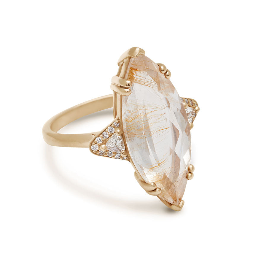 Angled view of a three stone engagement ring with a marquise shape copper rutilated quartz center stone with two white diamond trillion side stones with white diamond pave halos around them set in yellow gold.
