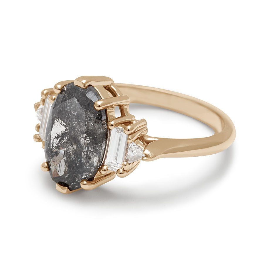 Angled view of a ring with an oval shaped painted black diamond center stone with one baguette white diamond and one white diamond trillion on either side set in yellow gold.