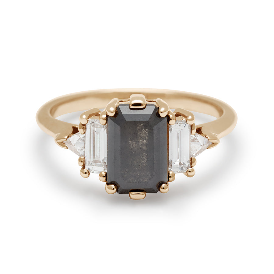 A ring with an emerald shaped black diamond center stone with one white diamond baguette and one white diamond trillion on either side set in yellow gold.