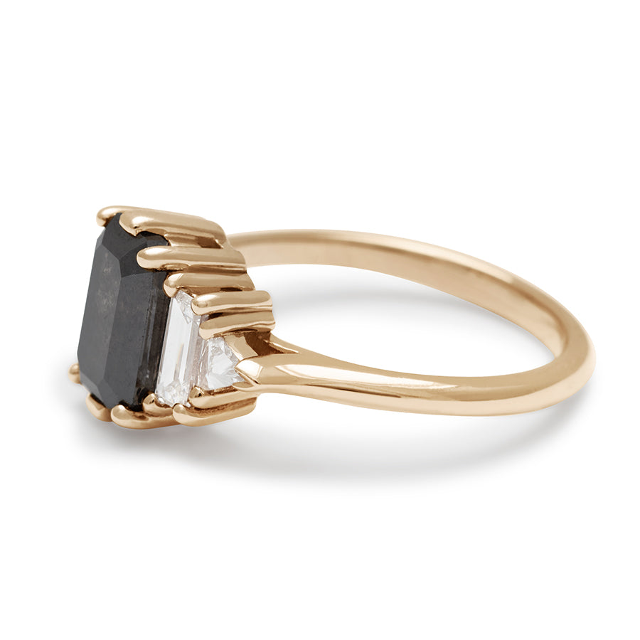 Angled view of a ring with an emerald shaped black diamond center stone with one white diamond baguette and one white diamond trillion on either side set in yellow gold.