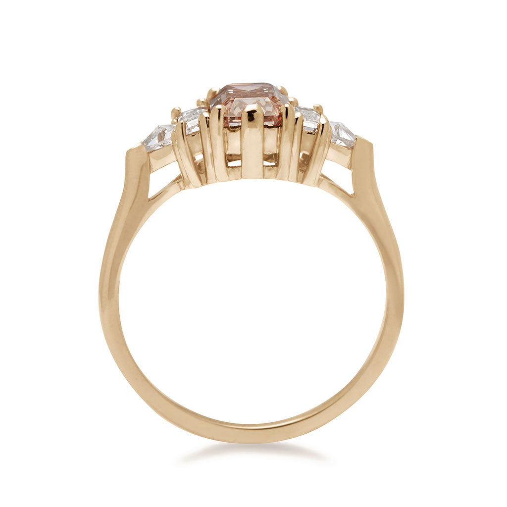 Side view of a ring with an emerald cut and shaped champagne diamond center stone with one baguette white diamond and one white diamond trillion on either side set in yellow gold.