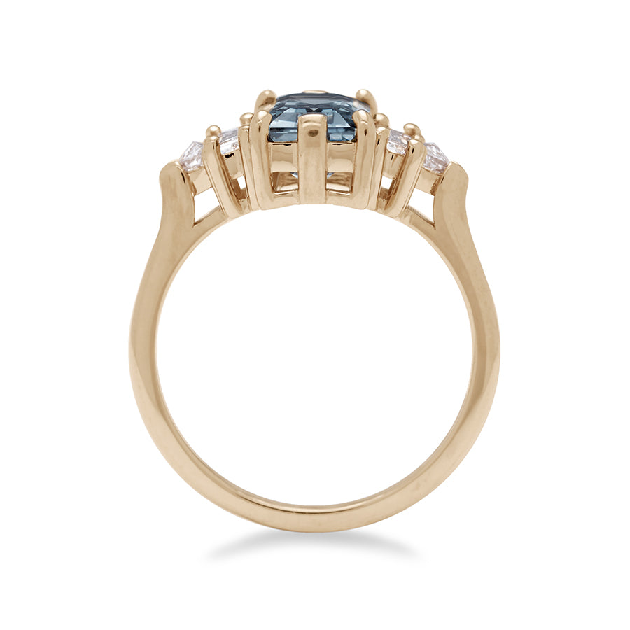 Side view of a ring with an emerald cut and shaped grey spinel center stone with one baguette white diamond and one white diamond trillion on either side set in yellow gold.