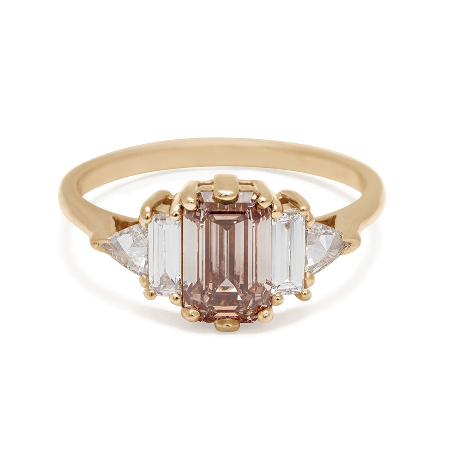 A ring with an emerald cut and shaped champagne diamond center stone with one baguette white diamond and one white diamond trillion on either side set in yellow gold.