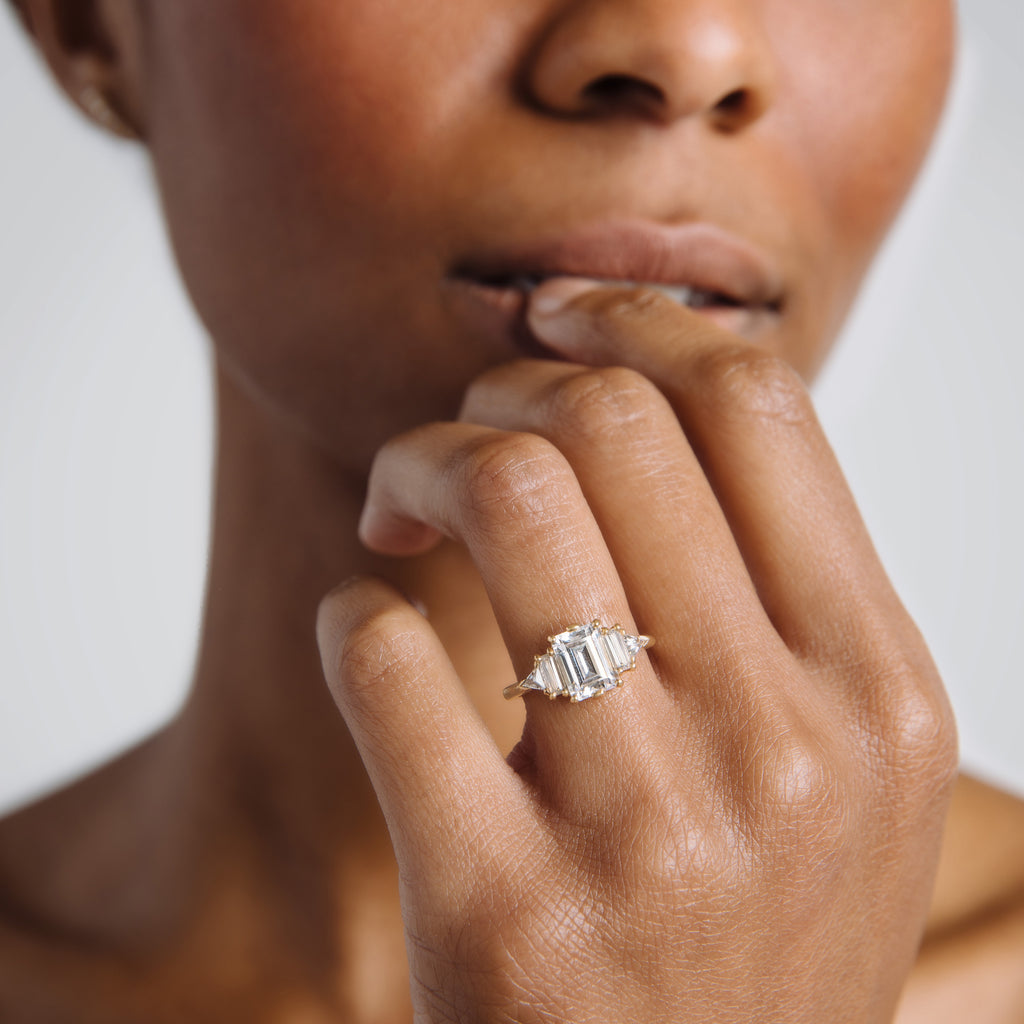A ring with an emerald cut and shaped 1.50ct white diamond center stone with one baguette white diamond and one white diamond trillion on either side set in yellow gold on a darker-complexion left hand ring finger., White Gold, Rose Gold, Yellow Gold