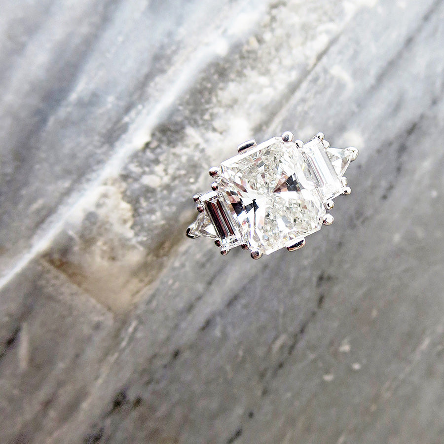 A ring with a brilliant cut and emerald shaped white diamond center stone with one baguette white diamond and one white diamond trillion on either side set in white gold on a grey marble stone.