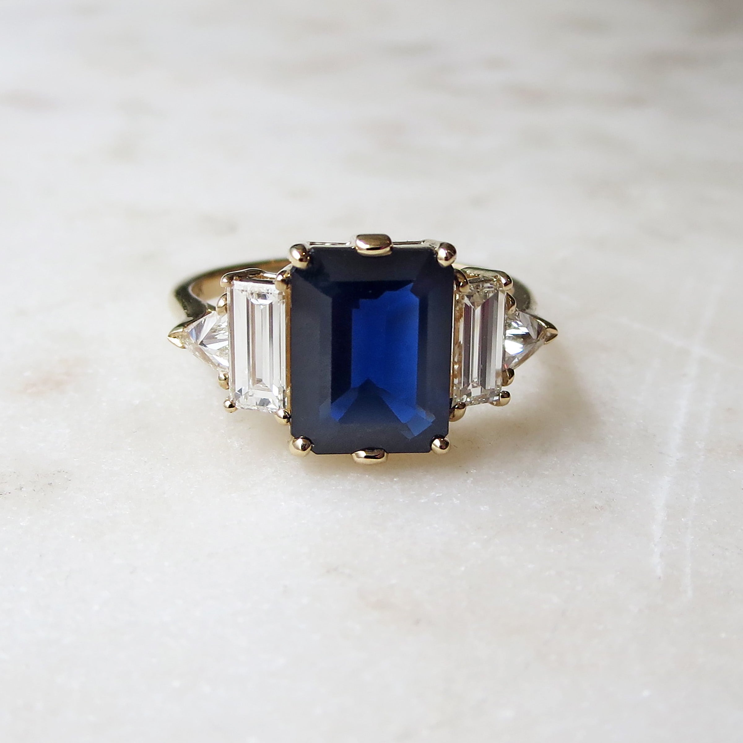 A ring with an emerald shaped blue sapphire center stone with one baguette white diamond and one white diamond trillion on either side set in yellow gold on a grey background.