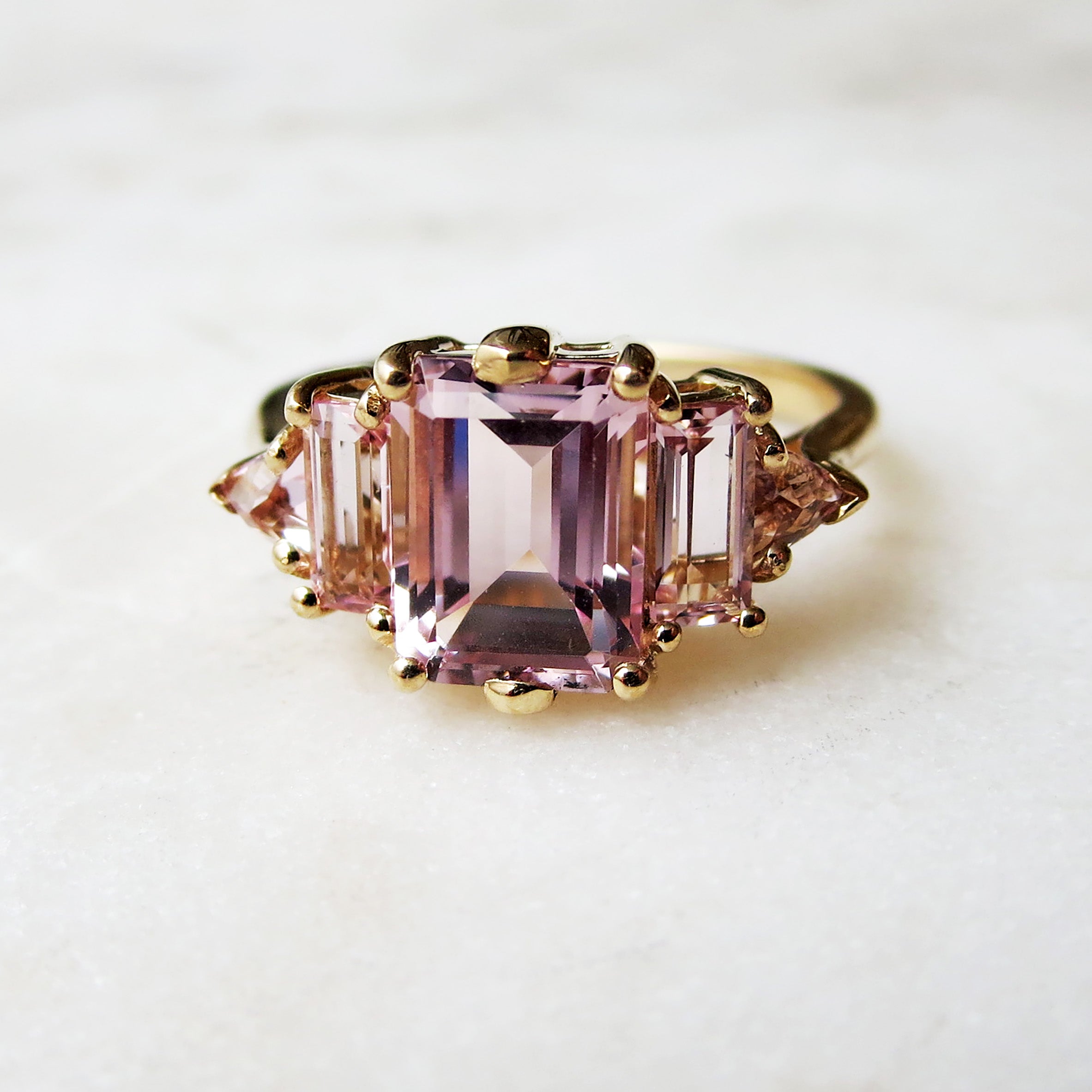 A ring with an emerald cut and shaped pink morganite center stone with one baguette pink morganite and one pink morganite trillion on either side set in yellow gold on a grey background.