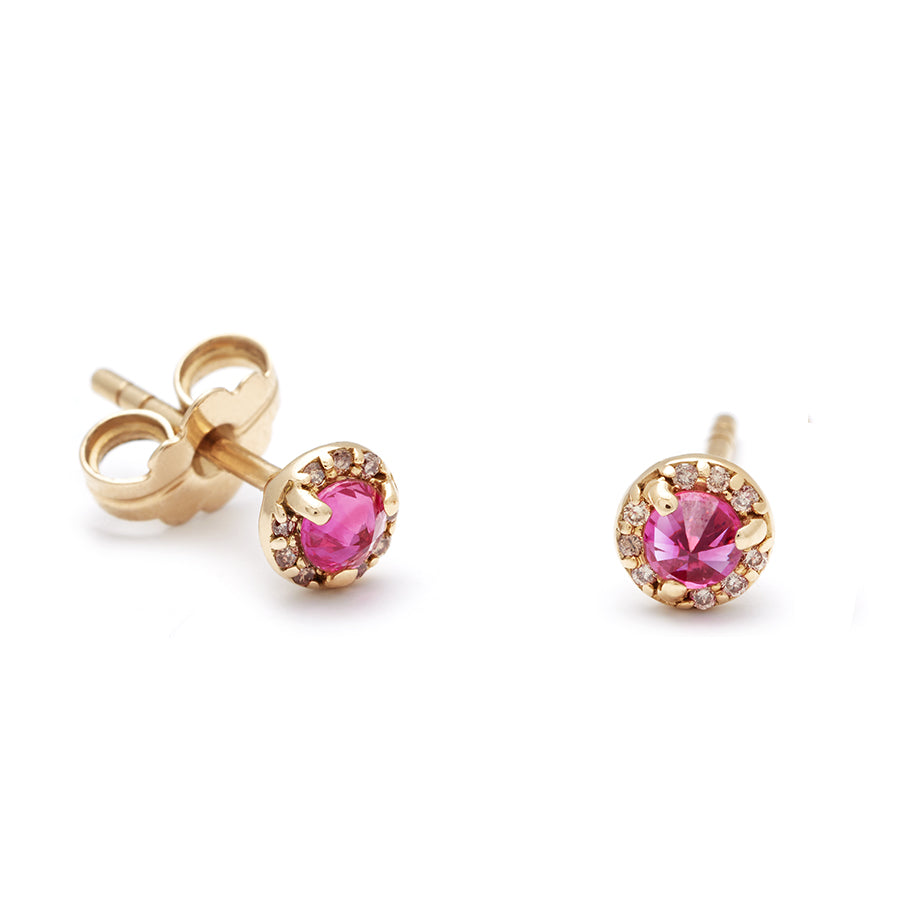 A pair of tiny ruby center stone earrings with a halo of champagne diamond set in yellow gold one earring angled.