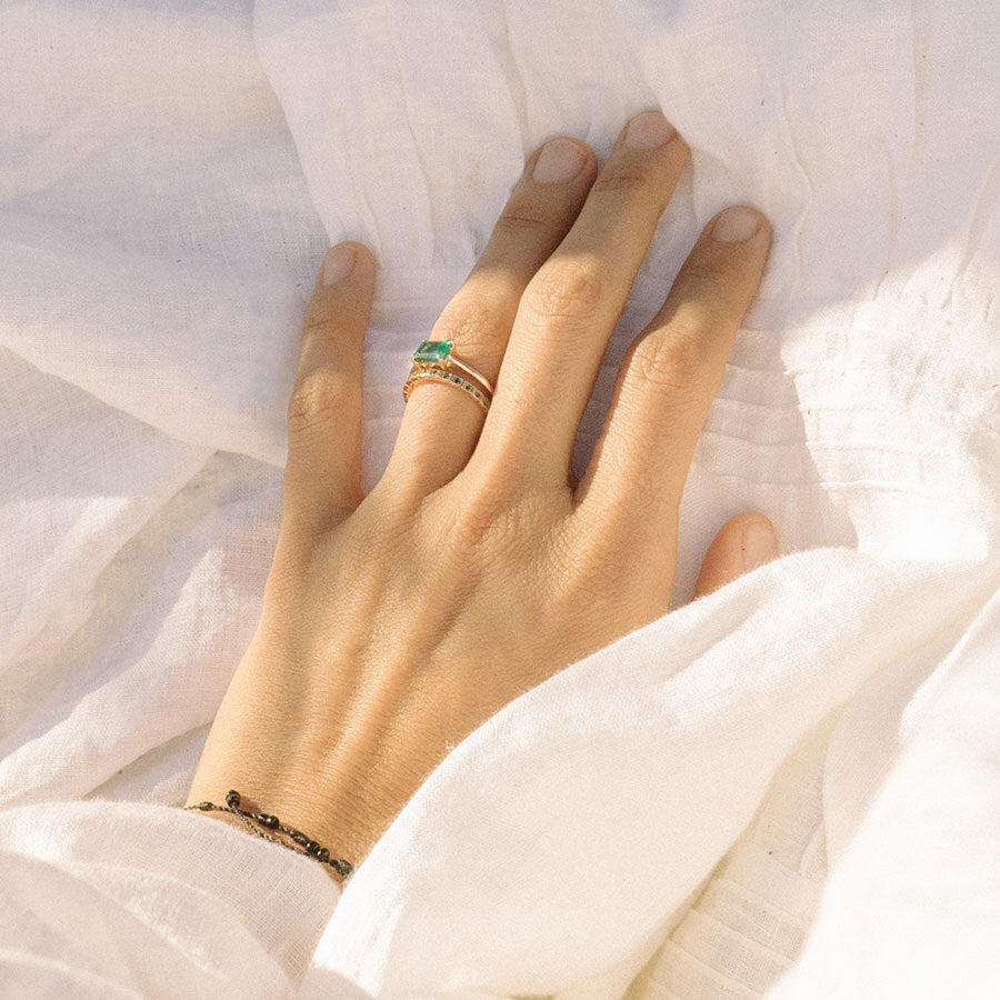 A straight band with emeralds set in eternity around the band set in yellow gold stacked beneath a solitaire east/west set emerald stone ring on a light-complexion hand on top of a right sheet.