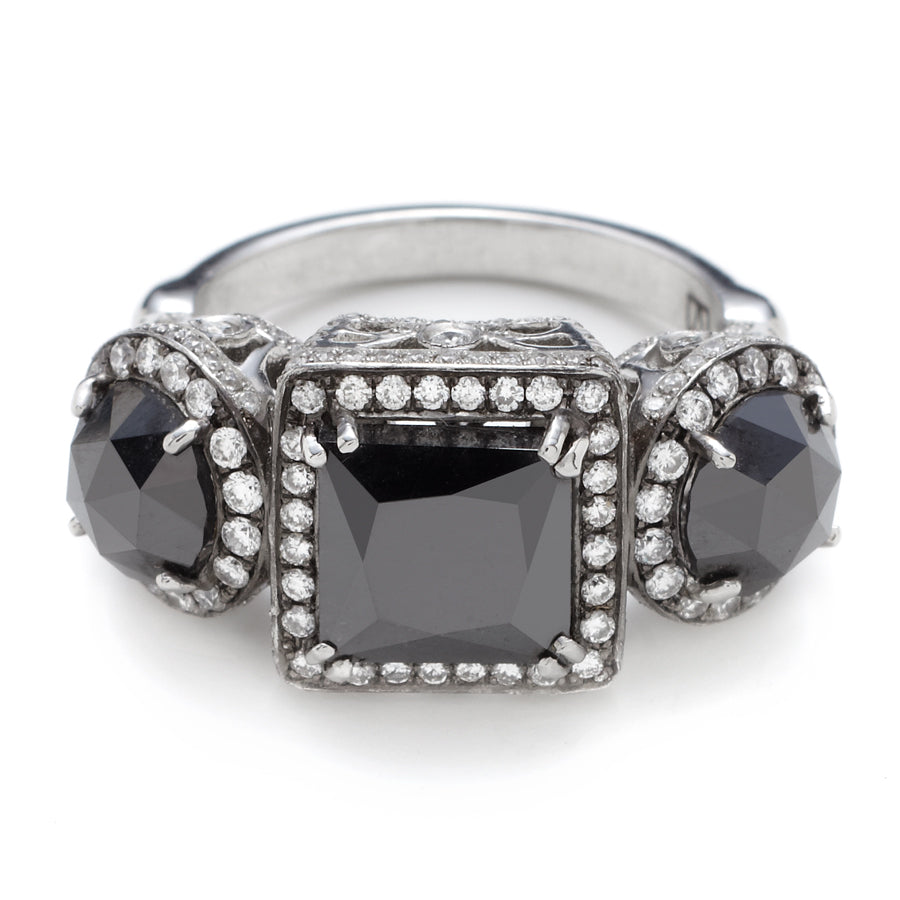 A three stone ring with an emerald cut and emerald shape black diamond center stone with two round black diamond side stones. All surrounded with white diamond pave halo set in platinum.