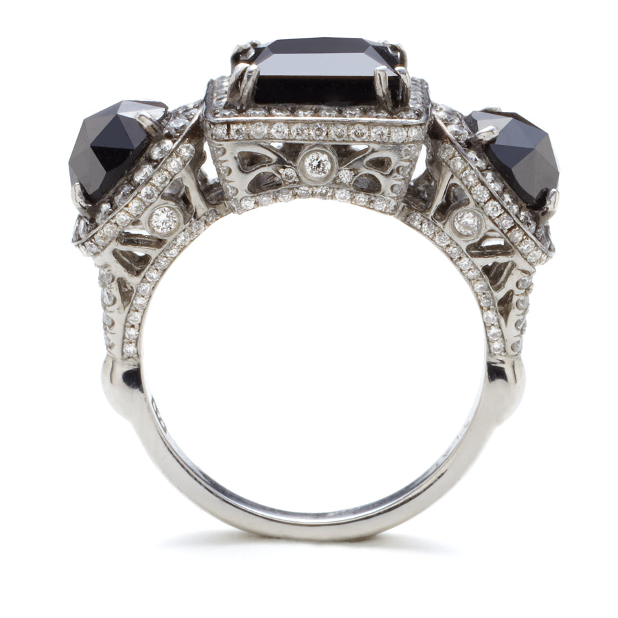 Side view of a three stone ring with an emerald cut and emerald shape black diamond center stone with two round black diamond side stones. All surrounded with white diamond pave halo set in platinum.