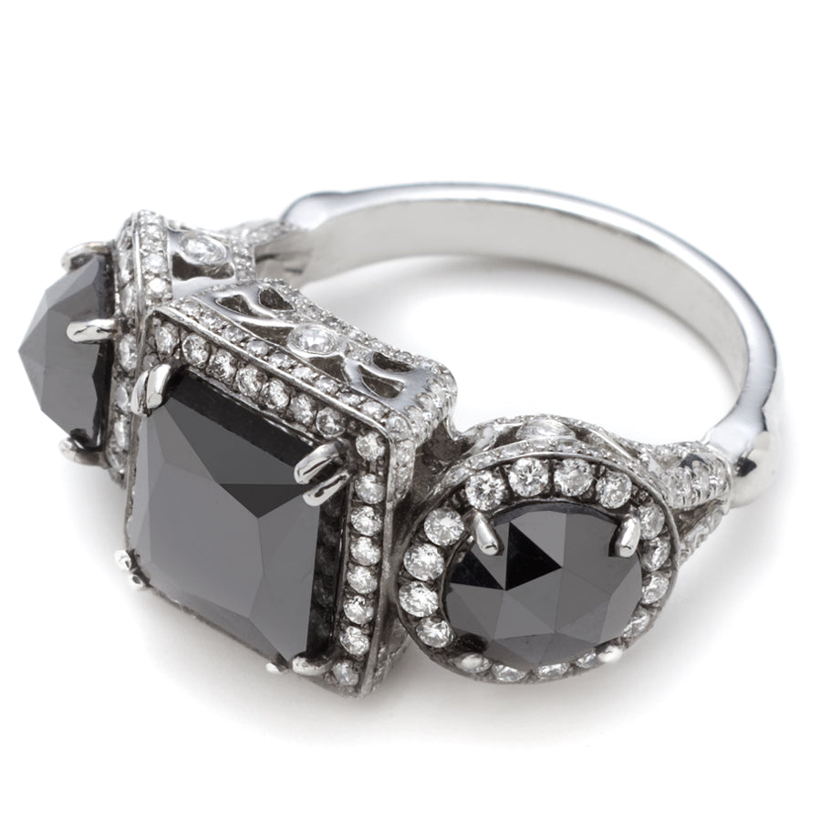 Angled view of a three stone ring with an emerald cut and emerald shape black diamond center stone with two round black diamond side stones. All surrounded with white diamond pave halo set in platinum.