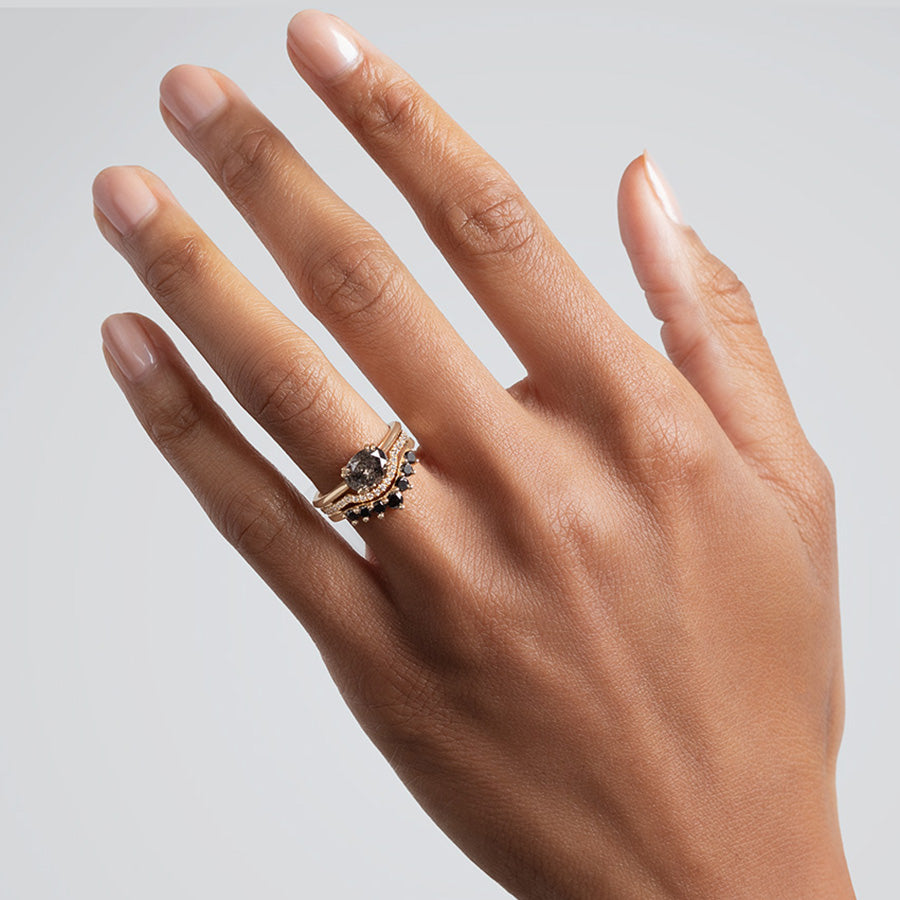 A solitaire grey diamond engagement ring stacked above a curved 7 black diamond band set in yellow gold on a medium-complexion model.