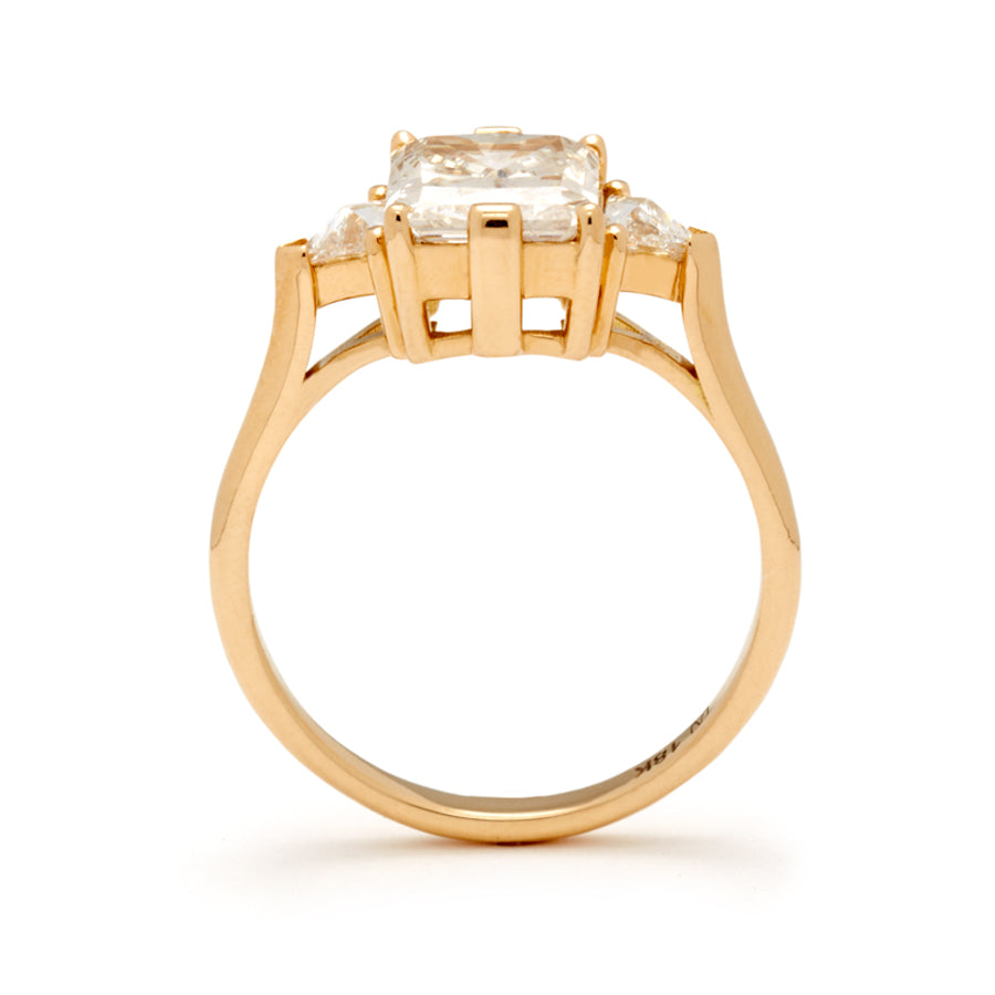Side view of a three stone engagement ring with an emerald shape champagne diamond center stone with two white diamond trillion side stones set in yellow gold.