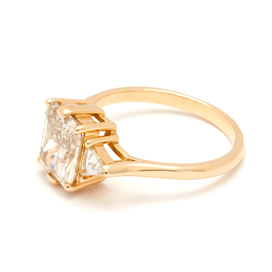 Angled view of a three stone engagement ring with an emerald shape champagne diamond center stone with two white diamond trillion side stones set in yellow gold.