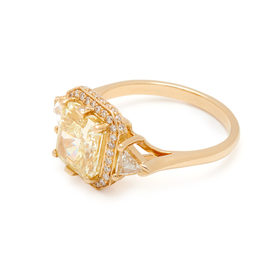 Angled view of a three stone engagement ring with an radiant cut yellow diamond center stone surrounded by a white diamond pave halo with two white diamond trillion side stones set in yellow gold.