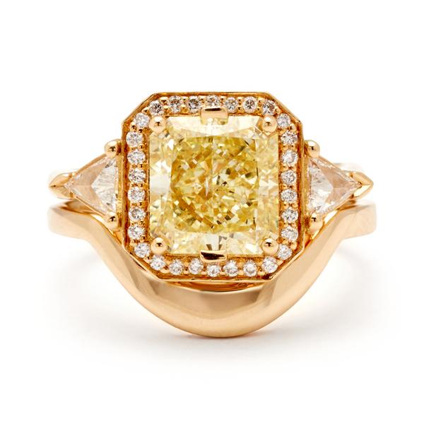 A three stone engagement ring with an radiant cut yellow diamond center stone surrounded by a white diamond pave halo with two white diamond trillion side stones set in yellow gold above a tapered curve solid gold band.