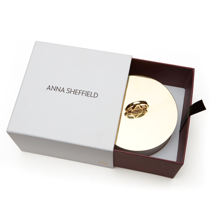 A white candle with a gold lid and the handle is shaped in the logo of Anna Sheffield in the box.