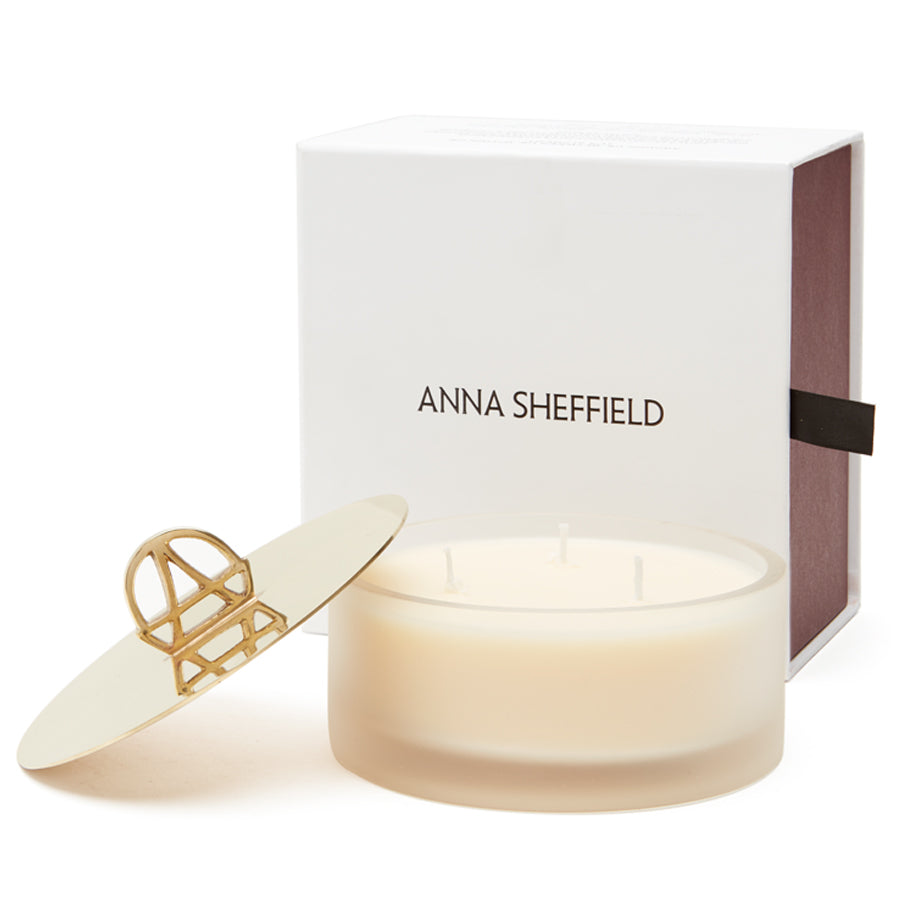 A white candle with a gold lid and the handle is shaped in the logo of Anna Sheffield with the lid open and propped against the candle box in the back of the image.