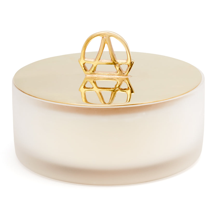 A white candle with a gold lid and the handle is shaped in the logo of Anna Sheffield.