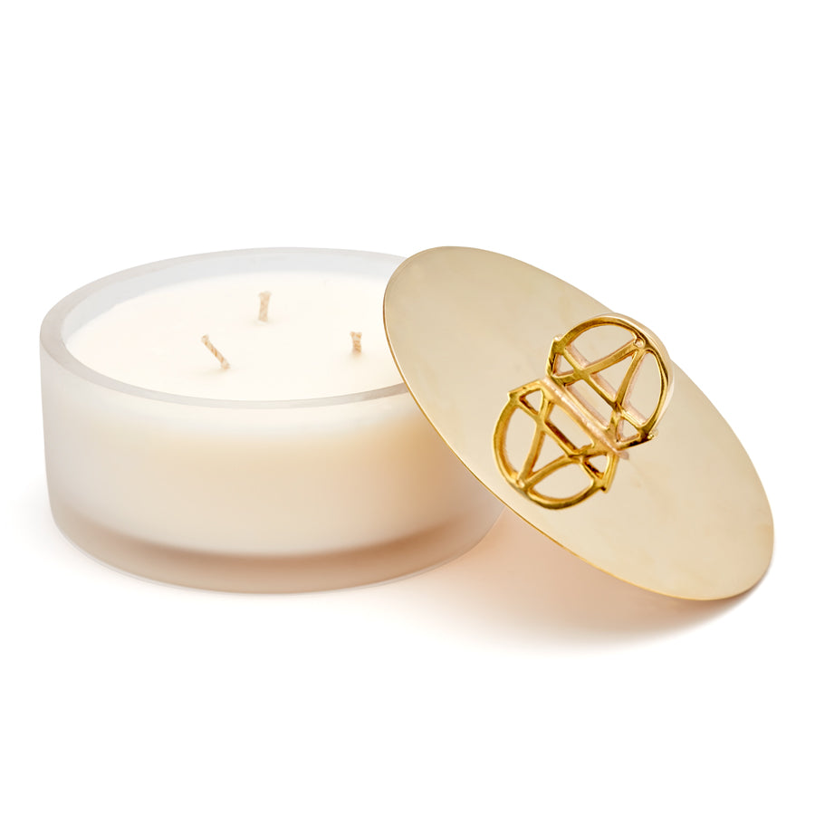 A white candle with a gold lid and the handle is shaped in the logo of Anna Sheffield with the lid open and propped against the candle.