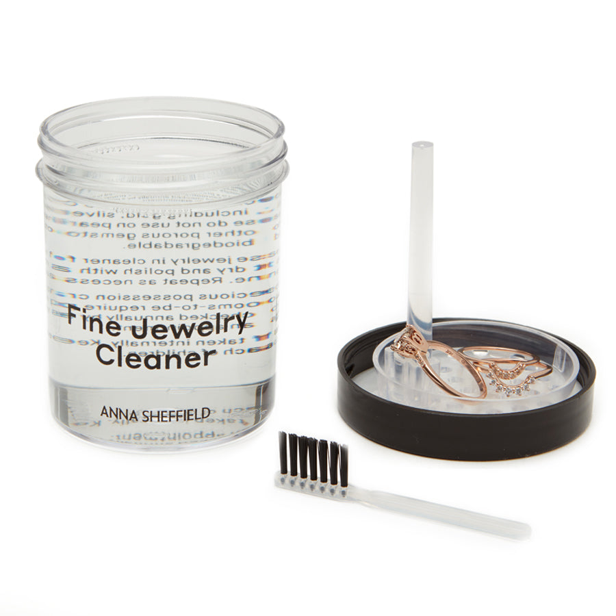 "Anna Sheffield Fine Jewelry Cleaner" in a small jar with clear liquid with a brush on the side with two diamond bands and an engagement ring.