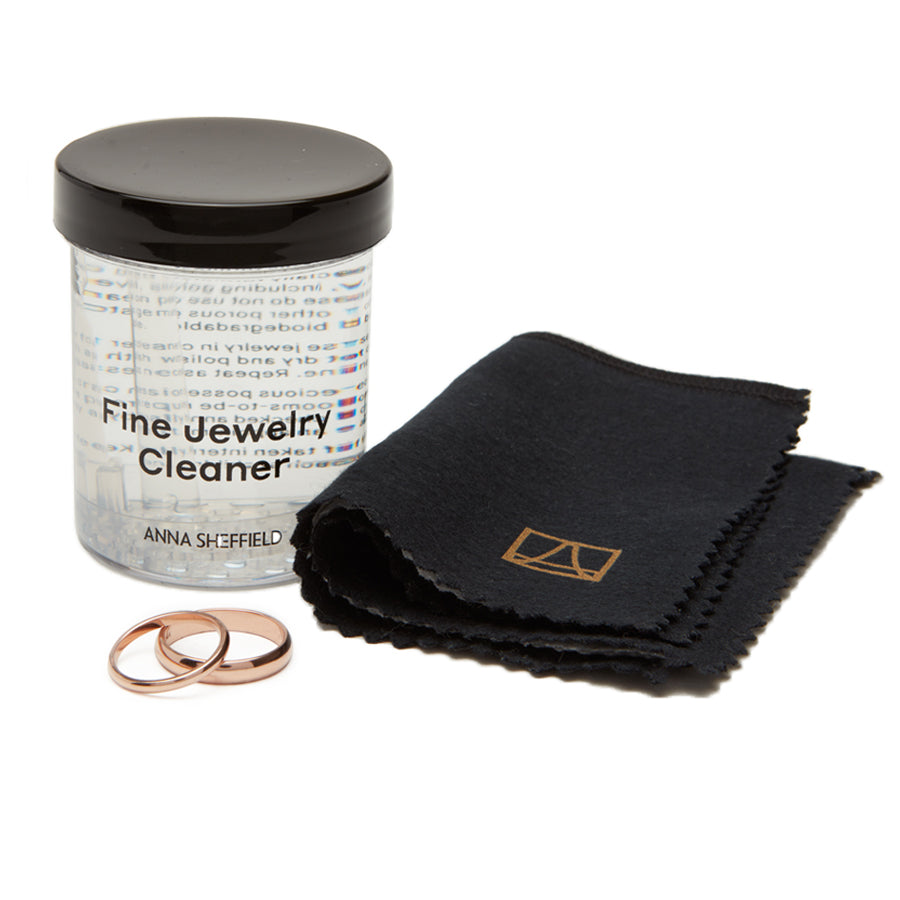 "Anna Sheffield Fine Jewelry Cleaner" in a small jar with clear liquid with a jewelry cloth next to it and two solid gold bands.