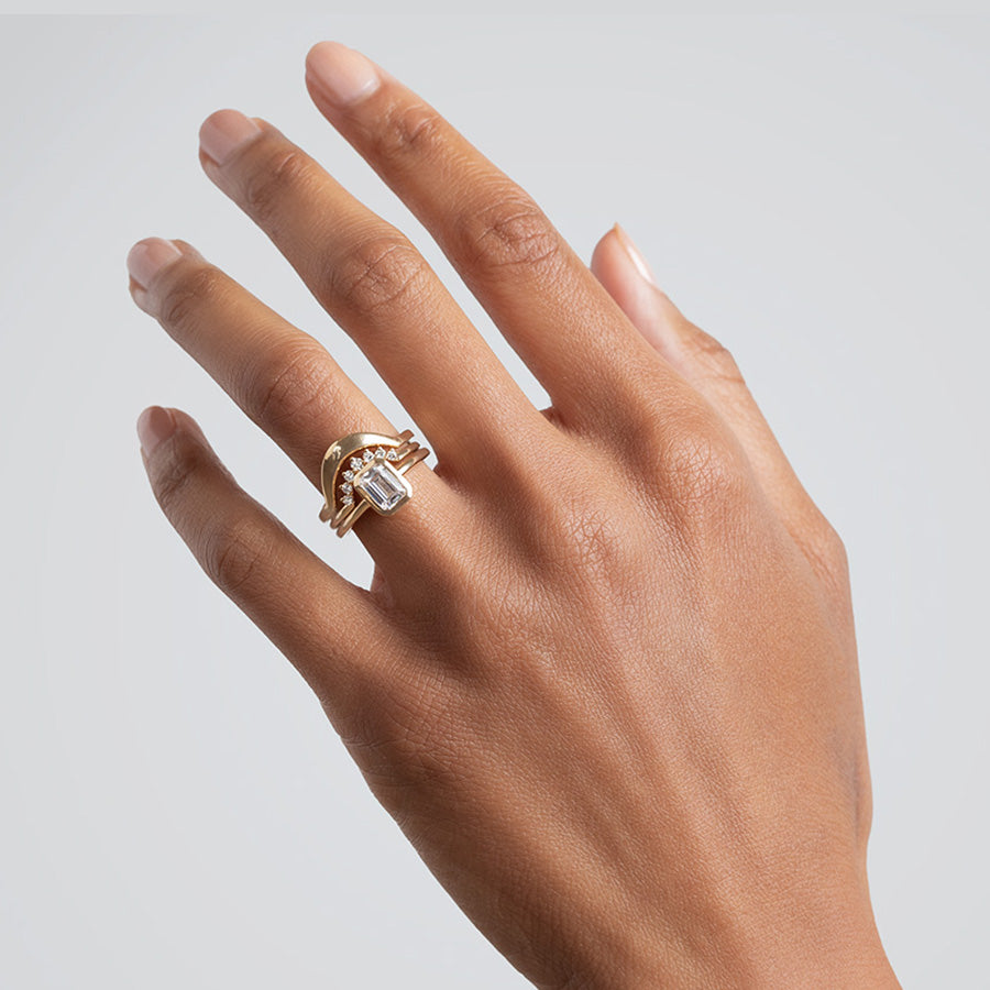 A elongated emerald cut bezel set white diamond set in yellow gold stacked below a 7 white diamond curve band and a tapered solid curve band in yellow gold on a medium-complexion ring finger., White Gold, Rose Gold, Yellow Gold
