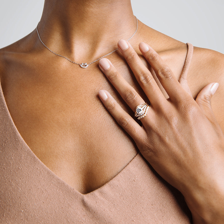 A solitaire ring with a round shape white diamond with white diamond pave on either side set in yellow gold inbetween three diamond dusted curve bands on a darker-complextion hand that is touching a neck that is wearing an arc necklace with a white diamond baguette., White Gold, Rose Gold, Yellow Gold