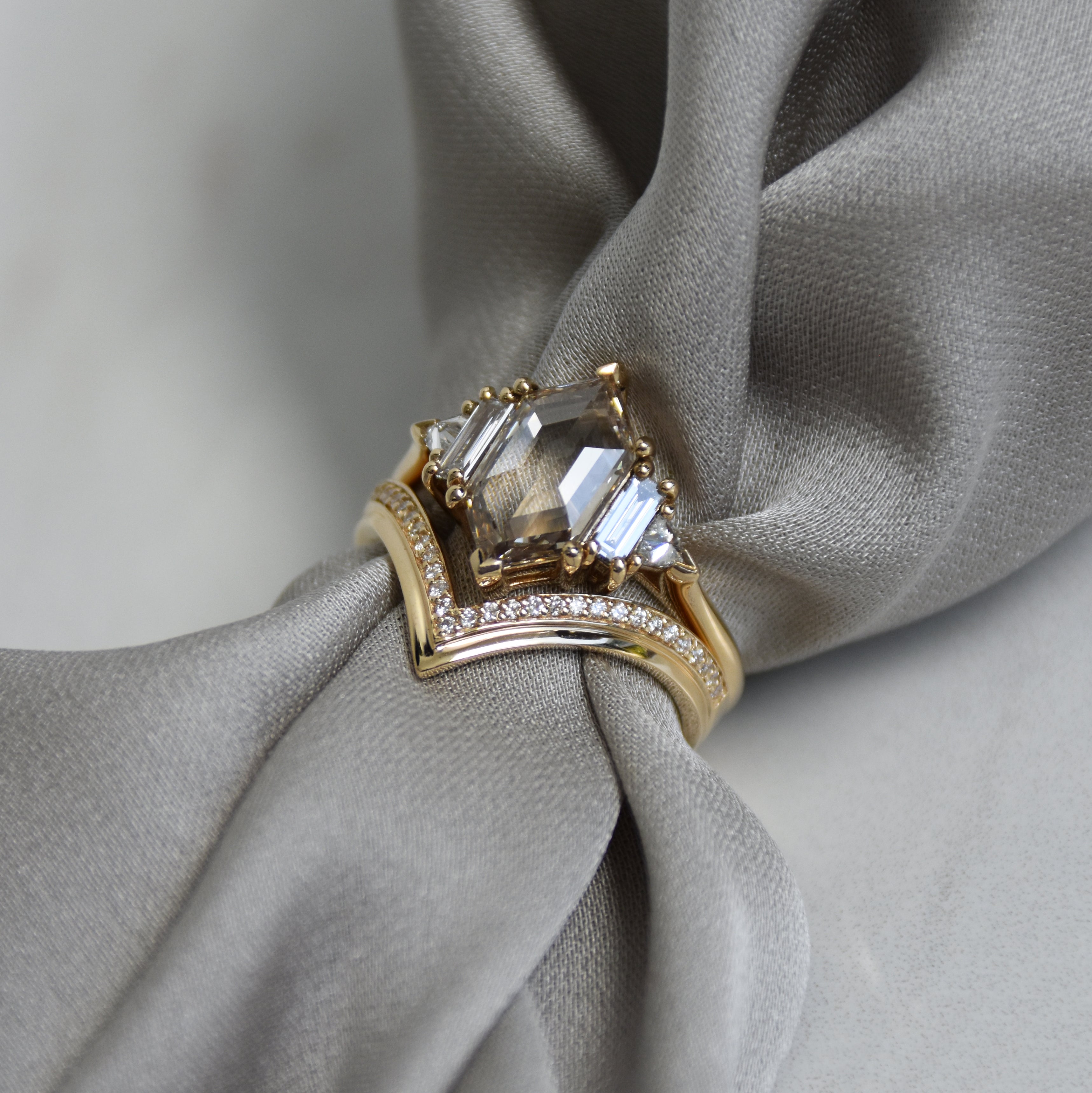 A ring with a hexagon shaped champange diamond center stone with one baguette white diamond and one white diamond trillion on either side set in yellow gold stacked above a chrysalis shaped white diamond band on grey fabric.