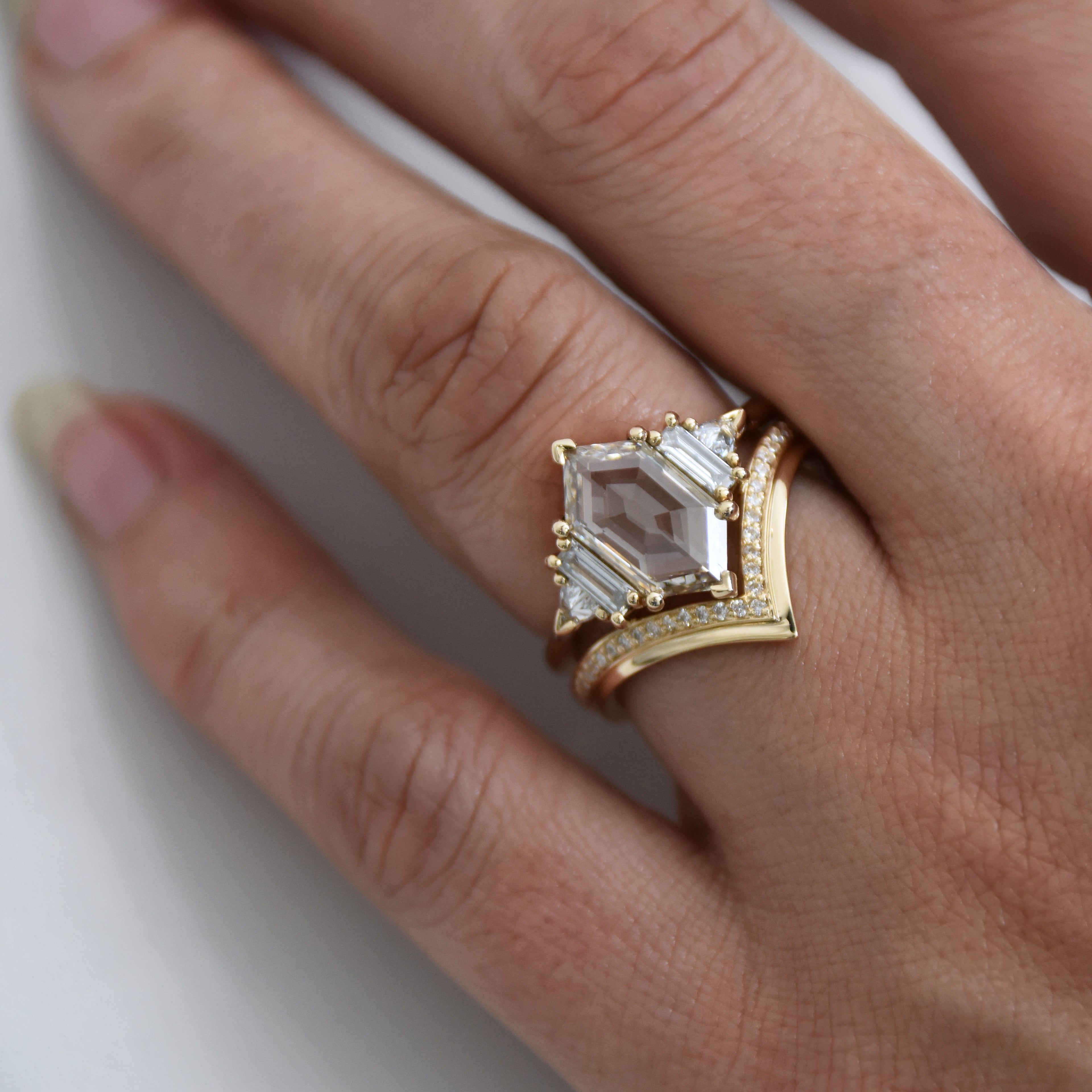 A ring with a hexagon shaped champange diamond center stone with one baguette white diamond and one white diamond trillion on either side set in yellow gold on a medium-complexion ring finger stacked above a chrysalis shaped white diamond band.