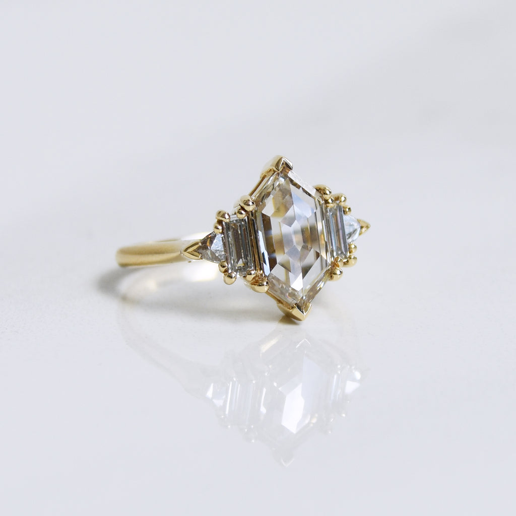 Angled view of a ring with a hexagon shaped champange diamond center stone with one baguette white diamond and one white diamond trillion on either side set in yellow gold on a grey background.