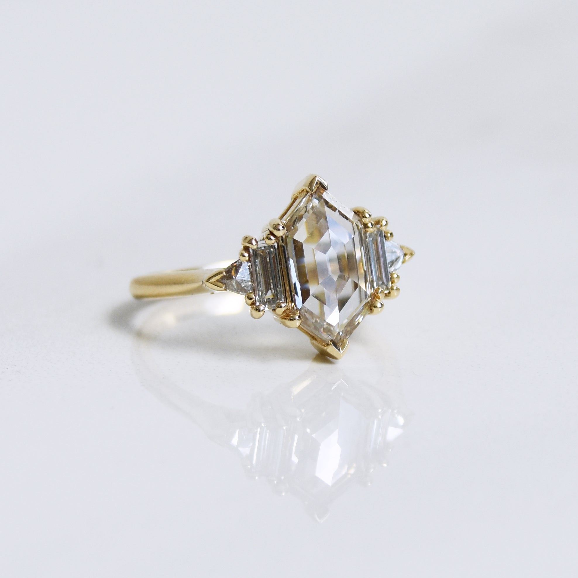 Angled view of a ring with a hexagon shaped champange diamond center stone with one baguette white diamond and one white diamond trillion on either side set in yellow gold on a grey background.