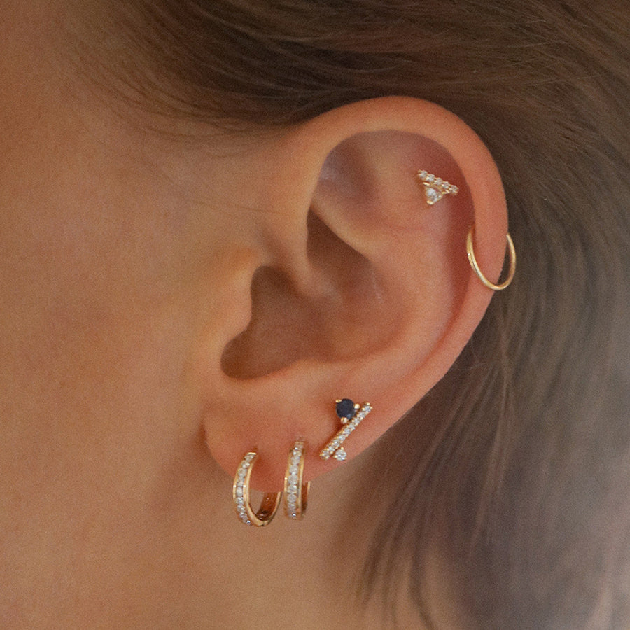 A single earring with a white trillion set above a line of white diamond pave set in yellow gold on the top of a light-complexion models ear at the base of the ear are two hoop earrings and a diamond dusted line stud., White Gold, Rose Gold, Yellow Gold