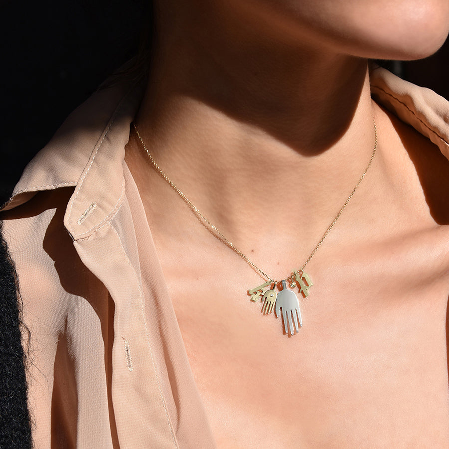 A large hand sterling sliver hand with a small yellow gold hand with a white diamond accent stone next to it on a yellow gold chain with two initial charms and a bezel charm on a light-complexion neck.