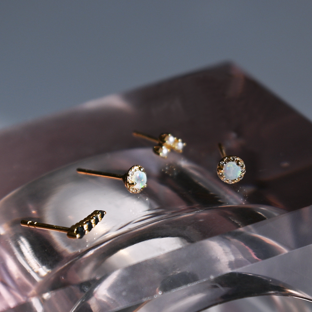 A single four black diamond stone pointe stud earring set in yellow gold on a clear table with two opal earrings and an arrow stud with three white diamonds., White Gold, Rose Gold, Yellow Gold