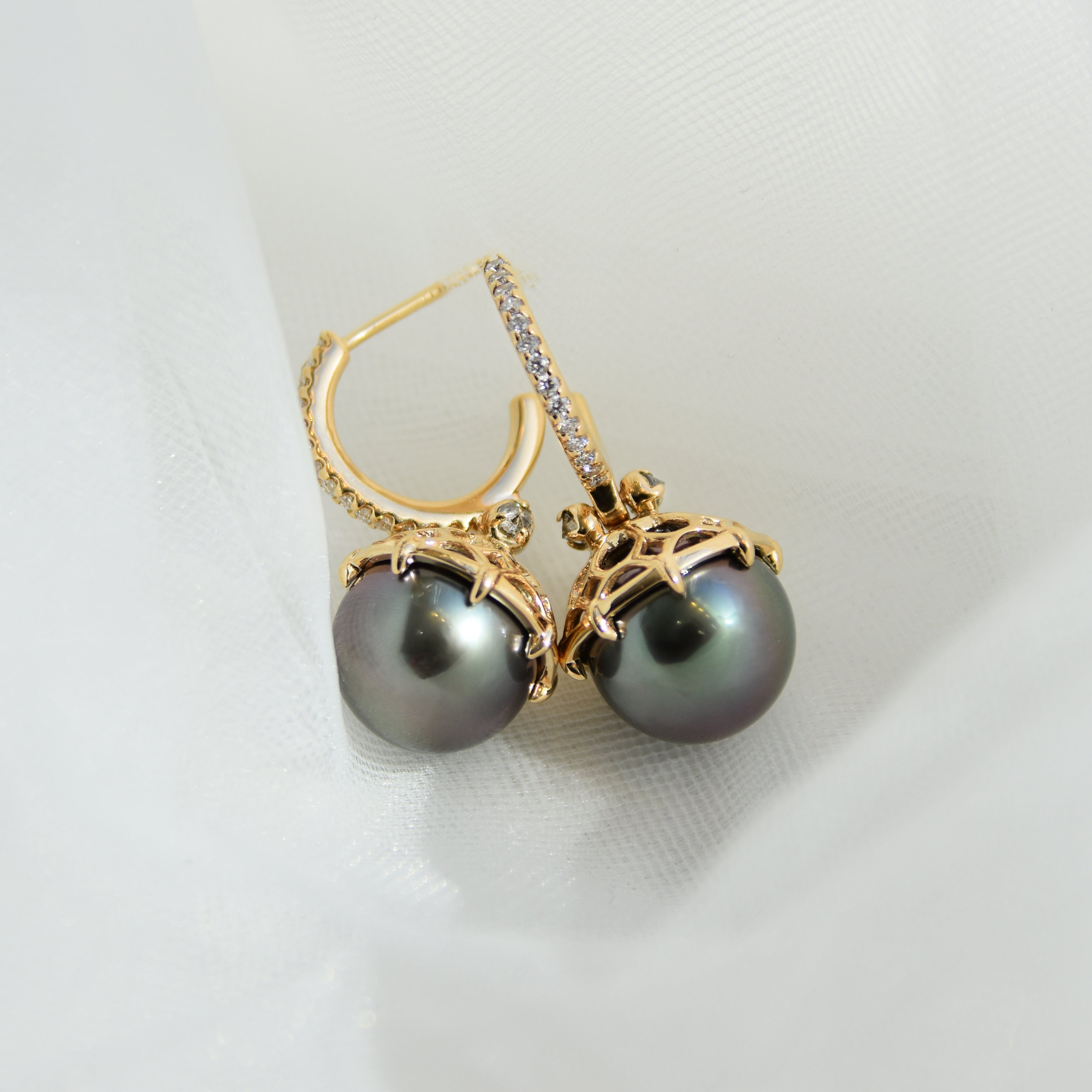 A pair of white diamond hoops with a dark pearl hanging from it with grey diamond accents on either side set in yellow gold in grey mesh.