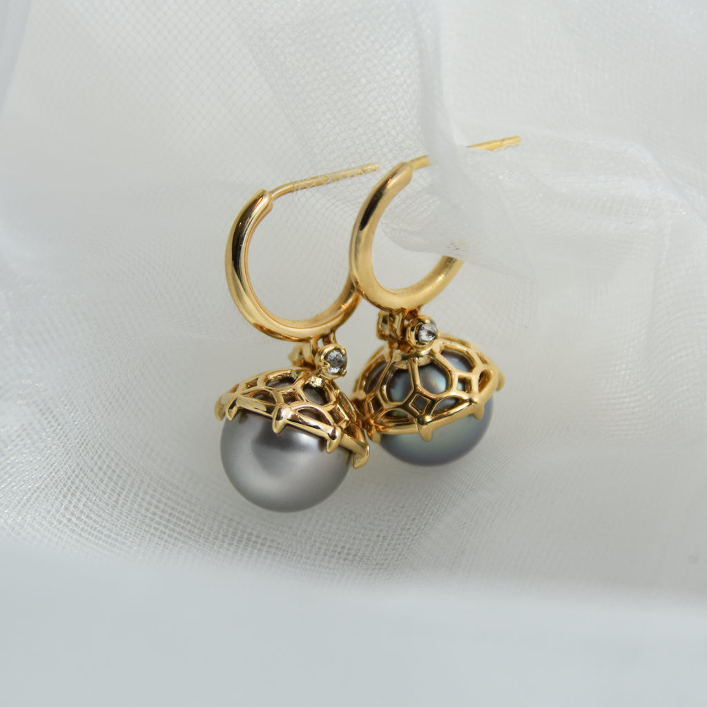 A pair of hoops with a light pearl hanging from it with grey diamond accents on either side set in yellow gold set in grey mesh.