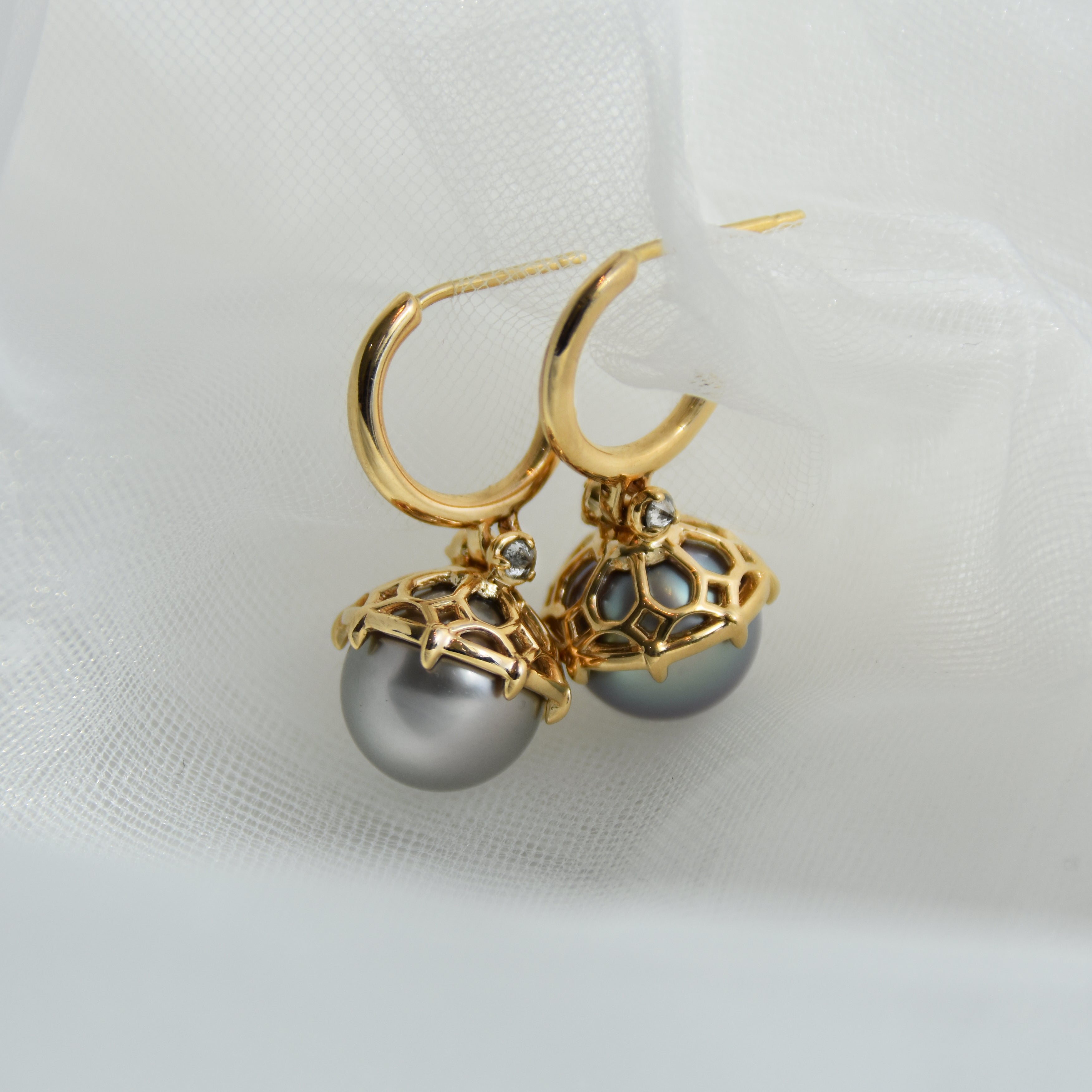 A pair of hoops with a light pearl hanging from it with grey diamond accents on either side set in yellow gold set in grey mesh.