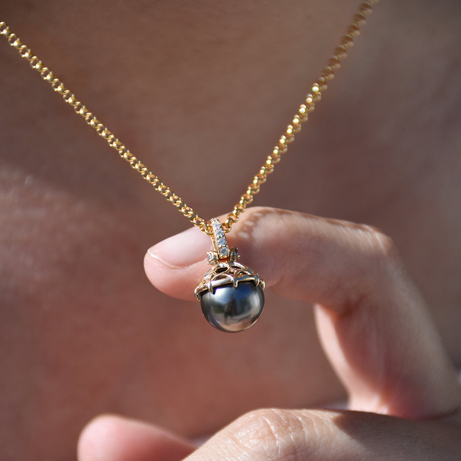 A one of a kind peacock pearl pendant with white diamond pave on the top and grey diamond accent stones on either side on a yellow gold chain with a medium-complexion finger holding it out from a model's neck.