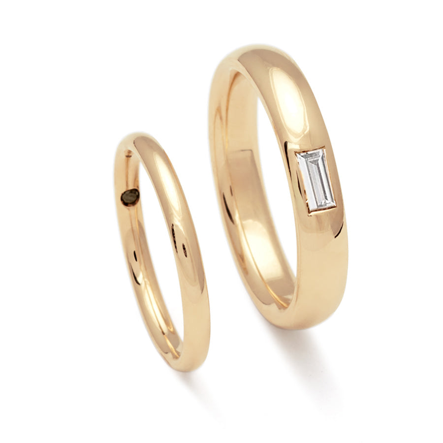 A 2mm classic round band set in yellow gold with a hidden black diamond next to a 4mm classic round band with a white diamond baguette set in yellow gold.
