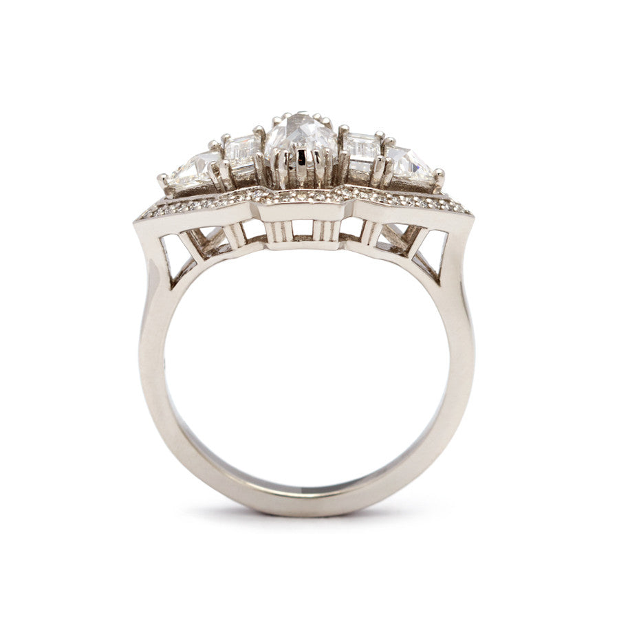 Side view of a ring with an emerald cut and shaped white diamond center stone with one baguette white diamond and one white diamond trillion on either side all encircled in white diamond pave halo set in platinum.